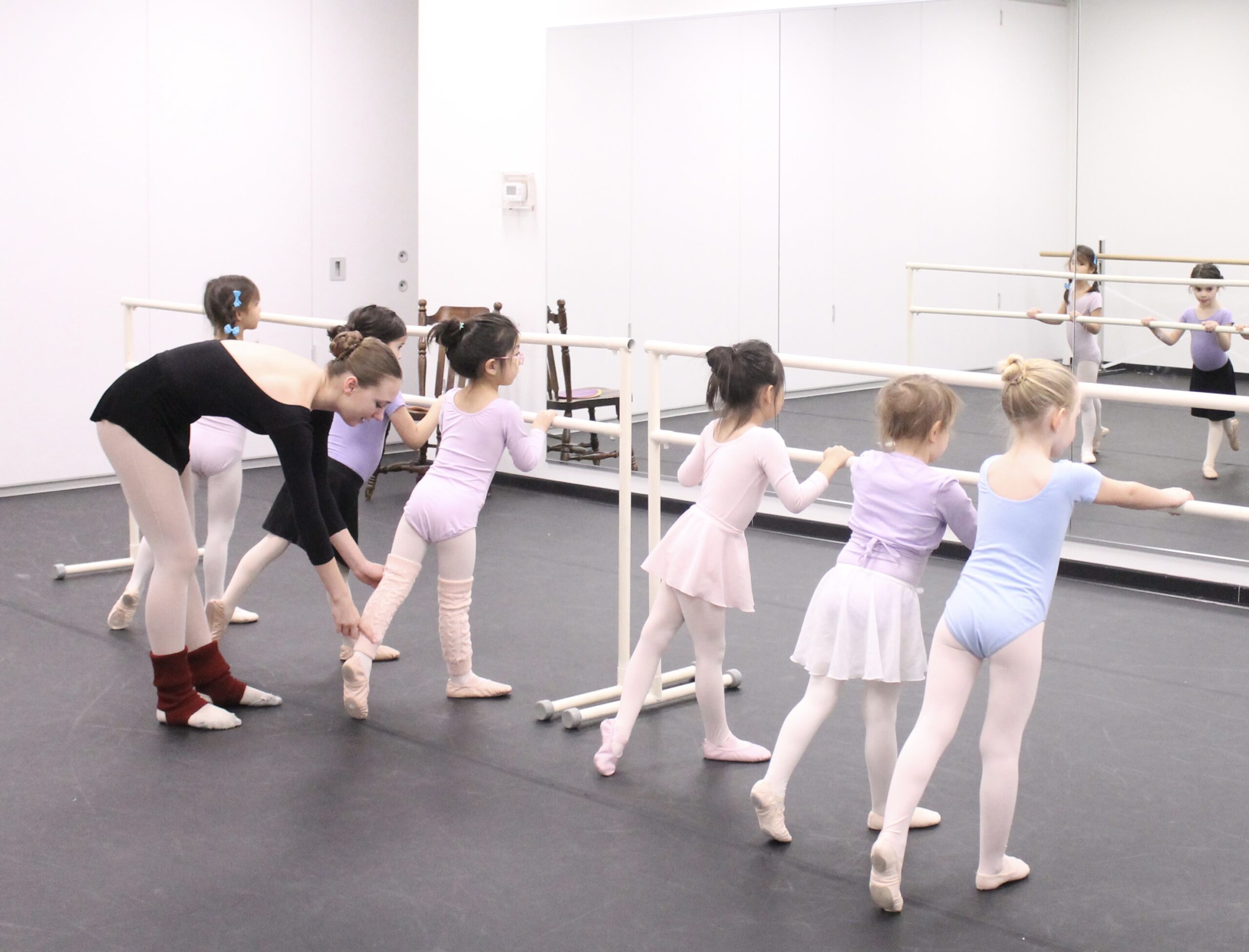 A teenager in a black leotard, pink tights and burgundy leg warmers corrects a young student's leg position while the student stands at barre with five classmates.