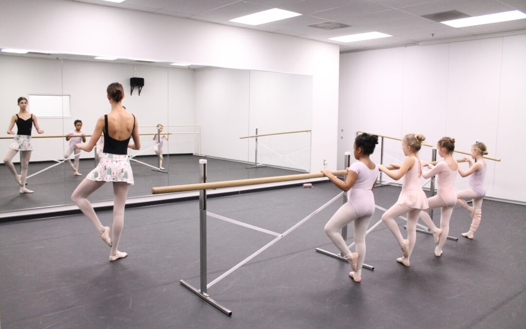 ballet teaching apprentice demonstrates coupe in front of a group of four little girls in pink and lavender leotards at the barre.