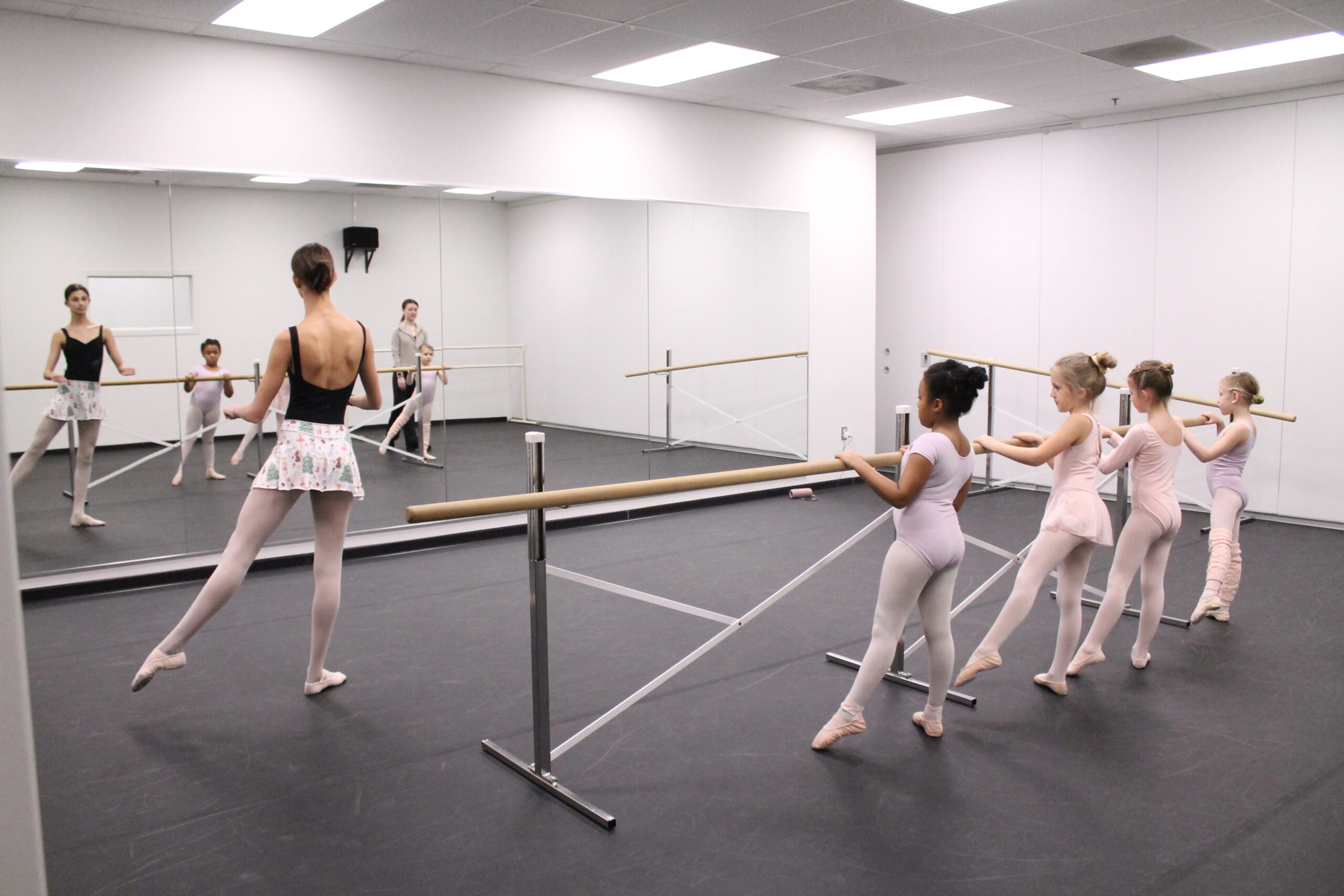 An advanced ballet student teaching apprentice holds her left leg in tendu in front of a group of four small girls mimicking her technique at the barre.